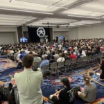 Image of a crowded room of attendees with a large DefCon banner at the front of the room.